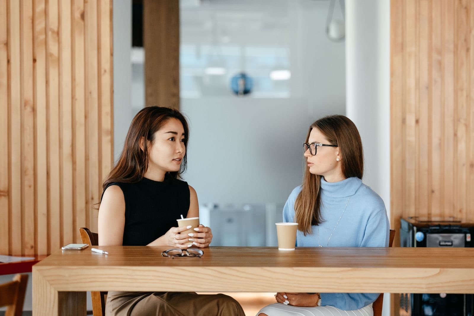 Twee collega's in gesprek bij de koffie aan een houten tafel op een lichte werkplek