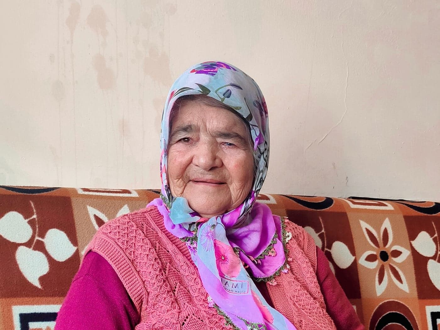 سيدة مسنة بغطاء رأس مزهر تجلس مبتسمة على الأريكة في غرفة معيشتها